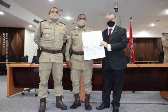 Desembargador Baltazar Miranda Saraiva segurando um certificado de “Título Policial Militar Padrão” e dois políciais militares fardados.